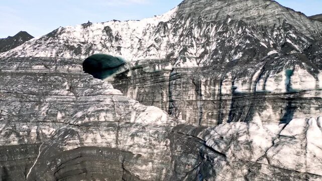 Cinematic aerial drone flight through Katla ice cave tunnel showing blue ice with black volcanic ash veins and green mossy mountains in Iceland 4K.