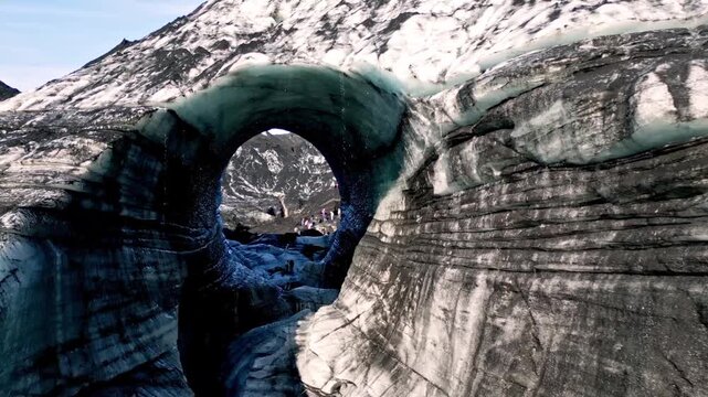 Cinematic aerial drone flight through Katla ice cave tunnel showing blue ice with black volcanic ash veins and green mossy mountains in Iceland 4K.