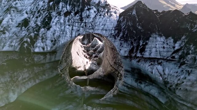 Cinematic aerial drone flight through Katla ice cave tunnel showing blue ice with black volcanic ash veins and green mossy mountains in Iceland 4K.