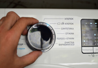 A hand turning the wheel of a washing machine - labels (cotton, synthetics, spin, spin) are visible nearby, and a display with indicators is visible on the right.