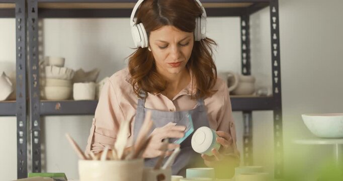 A woman in an apron and pink shirt shapes a turquoise ceramic mug in a bustling pottery studio, dust rising as she sands the piece with care, embodying craft, patience, and handmade artistry. Zoom in 