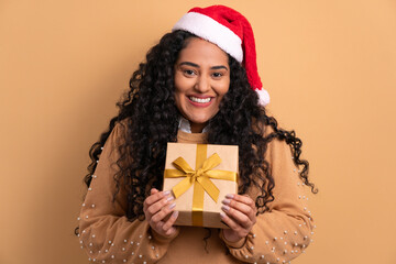 cheerful brazilian woman receiving gift box in beige background. christmas, holidays, festivity concept.
