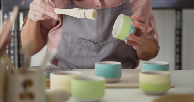 Female Potter in a pink shirt and gray apron, sending freshly made blue ceramic cup in a studio. A skilled potter sands and polishes a blue ceramic cup using a green sanding block in a workshop.