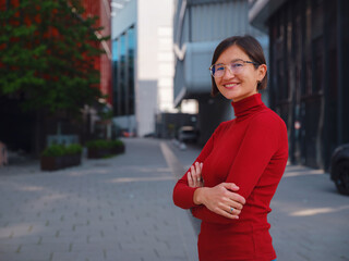 Woman in glasses and red turtleneck, dressed in business style, stands in center of Frankfurt, surrounded by modern buildings and urban life, embodying professionalism and confidence