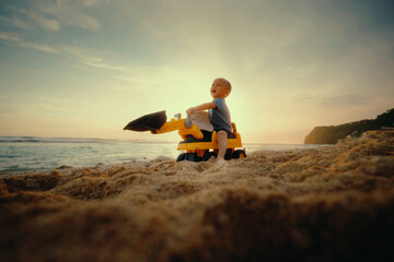 A young child happily enjoys playing with a toy excavator while exploring the sandy beach at a beautiful sunset