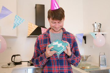 Young man receives gift at home, expressing surprise and happiness. Celebrating special occasion, he admires wrapped present in cozy, cheerful setting, capturing joyful and memorable moment