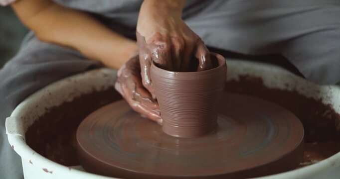 Close-up of a potter's muddy hands shaping brown clay on a spinning pottery wheel, creating a ceramic vessel. The image captures craftsmanship, focus, and creativity in a studio setting.