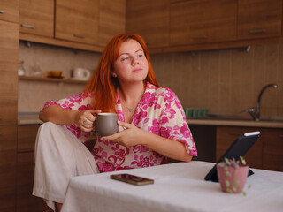 young woman working on tablet at home kitchen. A real female freelancer uses computer to work remotely from home with cup of morning coffee.