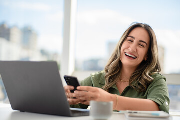 Laughing Latin businesswoman in her 30s wearing green shirt checking smartphone while working with laptop in bright office with urban skyline view and coffee mug on white desk