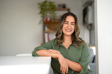 Smiling Latin businesswoman in green button-up shirt leaning on white table in cozy home office with wooden bookshelf and houseplant in background during daytime