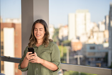 Cheerful businesswoman in green button-up shirt checking smartphone while standing on modern balcony overlooking urban cityscape 