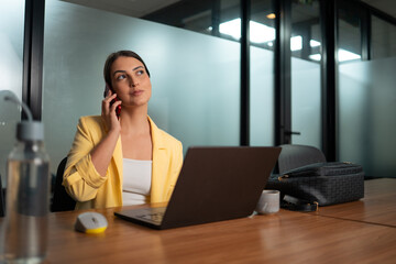 Young professional woman in yellow blazer on business phone call working on laptop in modern office