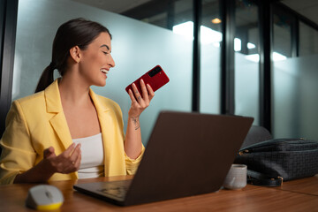 Professional woman in yellow blazer sending voice message on smartphone in modern corporate office workspace