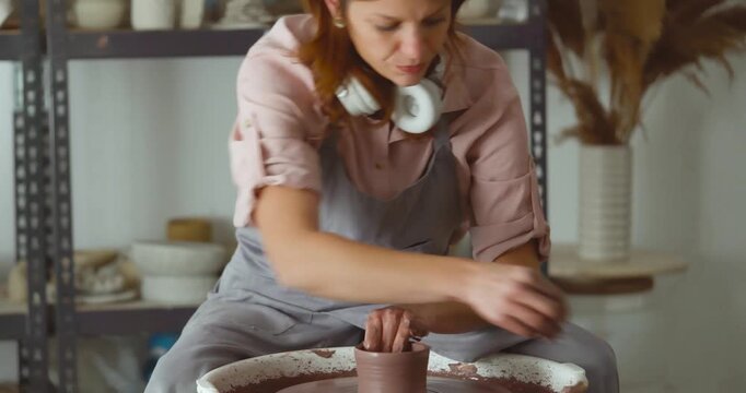 A female potter sits at a pottery wheel, hands kneading soft clay. In a well-lit studio, she concentrates on shaping a ceramic form, embodying craft, creativity, and handmade art. Tilt shoft down