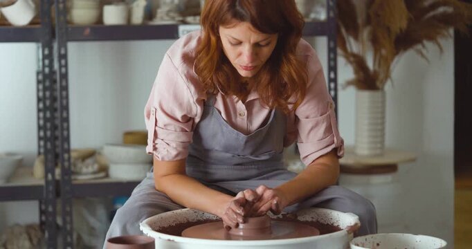 A dedicated potter shapes a clay piece on a pottery wheel in a busy studio, surrounded by ceramics on shelves. The scene conveys concentration and the joy of handmade craft. Static shot