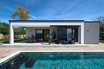 Modern modular house backyard featuring a rectangular swimming pool, large patio with wooden sun loungers, and umbrellas against a blue sky. Contemporary prefabricated home design