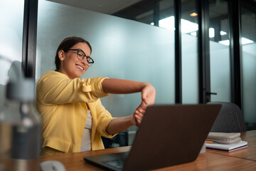 Professional woman in yellow blazer stretching arms during work break at desk in modern corporate office workspace