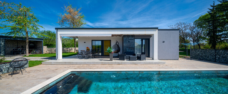 Modern modular house backyard featuring a rectangular swimming pool, large patio with wooden sun loungers, and umbrellas against a blue sky. Contemporary prefabricated home design