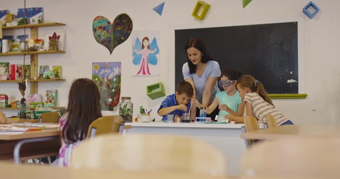 Female teacher guiding focused elementary students through a hands-on chemistry experiment using test tubes and liquids in a science classroom, with one student wearing safety goggles