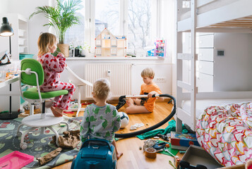 Children's room. Children elementary ages playing and cleaning in messy nursery. Boy vacuums the room while others siblings play. Child's playroom full of toys. Family at home.