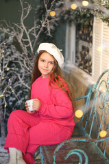 A little girl in a festive winter setting, sitting on a vintage bench and holding a warm cup. Child wearing a bright pink cozy outfit and a white beret. Christmas decorations. Holidays New Year concep