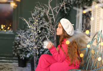 A little girl in a festive winter setting, sitting on a vintage bench and holding a warm cup. Child wearing a bright pink cozy outfit and a white beret. Christmas decorations. Holidays New Year concep