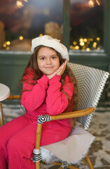 Adorable little girl in a festive winter setting, sitting on a vintage bench and holding a warm cup. Child wearing a bright pink cozy outfit and a white beret, smiling gently at the camera. Christmas