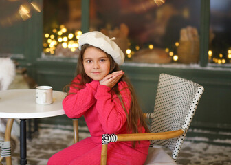 Adorable little girl in a festive winter setting, sitting on a vintage bench and holding a warm cup. Child wearing a bright pink cozy outfit and a white beret, smiling gently at the camera. Christmas