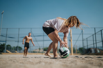 A woman kneels to retrieve a volleyball on a sunny beach court, with a shirt and shorts, while a man plays in the background. Outdoor sports, teamwork, fitness, and beach vibes.
