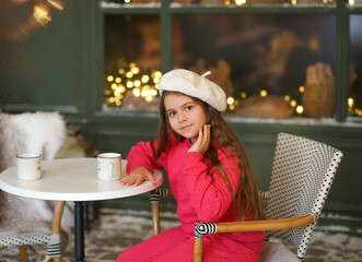 Adorable little girl in a festive winter setting, sitting on a vintage bench and holding a warm cup. Child wearing a bright pink cozy outfit and a white beret, smiling gently at the camera. Christmas