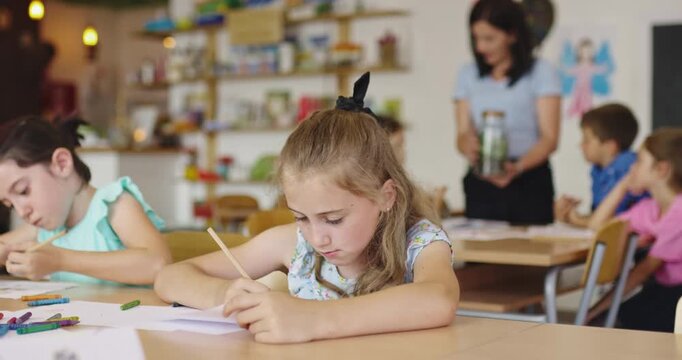 Focused elementary school girl writing at desk in classroom, doing homework or test, studying with classmates while teacher helps other children in the background.