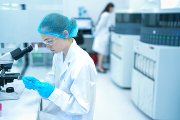 Fototapeta premium Young Caucasian female scientist in safety gear using micropipette and microscope in modern sterile lab. Focused research with colleague in background for medical science.