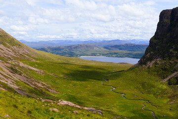 Naklejka premium Curved lush green valley with winding little creak in center and steep rocky and grassy cliffs each side in sunlight, lake and hilly mountains in the background, cloudy sky