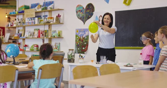 Smiling teacher conducting a science experiment with a balloon and lighter for excited elementary students in a classroom, while kids cover their ears in anticipation of the pop