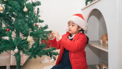 Young girl in Santa hat decorates Christmas tree with ornaments in cozy holiday home setting, capturing festive spirit and joyful tradition