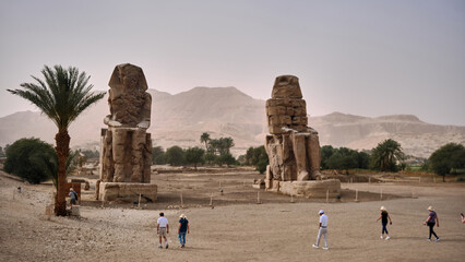 Wide view of ancient Colossi of Memnon standing in desert plain near Luxor, Egypt. Monumental stone statues rise against distant mountains and pale sky with visitors walking around archaeological site