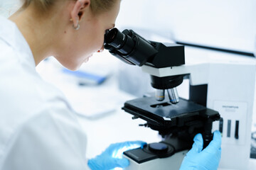 Close-up side view of Caucasian female scientist looking through microscope in bright lab. Young researcher in white coat and blue gloves performing medical analysis for biotechnology.