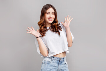 Beautiful teenage girl with wavy brown hair smiling and raising hands in playful gesture, posing in studio on gray background, young female fashion model in casual white top and denim