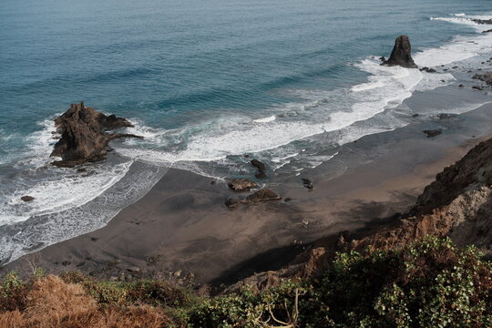Coastal landscape featuring black lava sand beach of Benijo, Tenerife, and rocky formations by the ocean, showcasing natural beauty and long waves perfect for surfing under a cloudy sky.