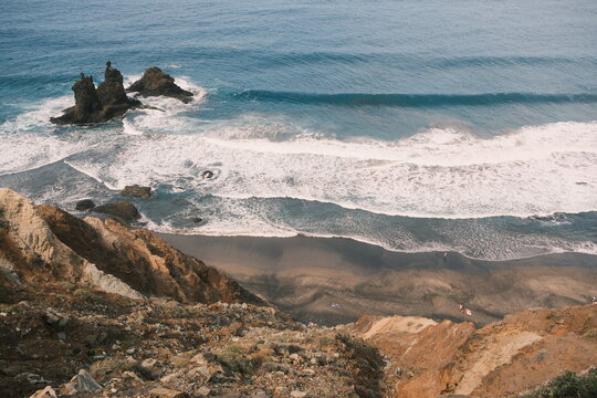 A scenic coastal view showing waves crashing against  rocky shores and the black sandy beach of Benijo, Tenerife, under a clear sky, perfect for nature and travel themes.