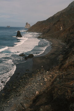 Rocky coastline with crashing waves and rugged cliffs under a cloudy sky, showcasing natural coastal beauty and ocean scenery of Benijo Beach.