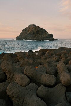 Rocky coastline with waves crashing against basalt formations and a small island in the distance under a soft sunset sky.