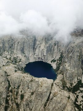 Aerial view of a deep blue mountain lake surrounded by rugged rocky cliffs and partially covered by low clouds.