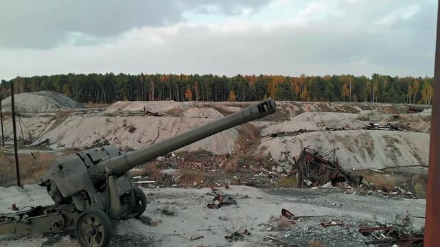 A weathered, deactivated artillery piece rests in a barren landscape near a colorful autumn forest