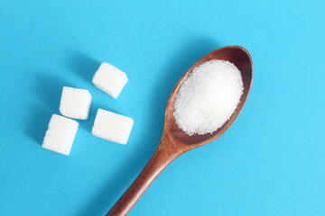 On a blue background there is loose and refined sugar lying on a wooden spoon.