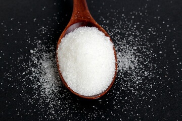 On a black background, there is white crumbly sugar lying on a wooden spoon.