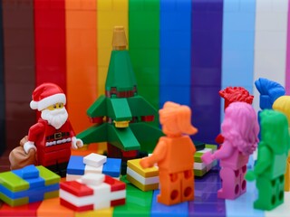 Naklejka premium Tambov, Russian Federation - October 19, 2025 Lego Santa Claus standing near Christmas tree with gifts in front of rainbow monochrome minifigures. Rainbow background