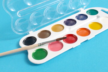 On a blue background lies a palette of watercolor paints and a brush.