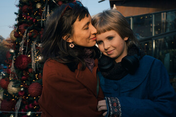smiling mother hugs teen daughter in winter city decorated by Christmas lights