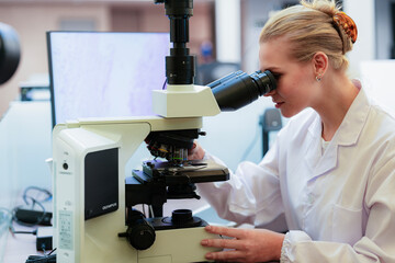 Smiling Caucasian female scientist looking through microscope in modern lab. Researcher analyzing histology slide with digital pathology monitor in background.